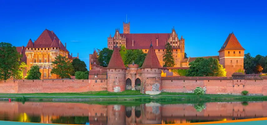 Malbork Castle seen from across the Nogat River at dusk, its red brick walls and towers reflected in the water
