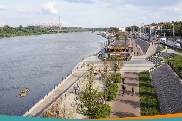 Warsaw's Vistula boulevards on a summer day with cyclists and pedestrians, the National Stadium and Świętokrzyski Bridge in the background